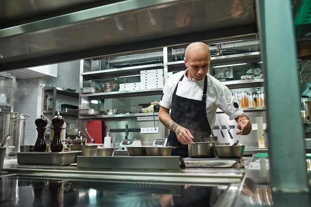 concentrated young chef in black apron is cooking at his modern restaurant kitchen