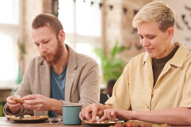 Concentrated young couple in casual outfits sitting at table and decorating cookie together for hall