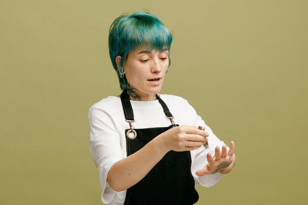 Concentrated young female barber wearing uniform cutting her nails with nail clippers isolated on olive green background