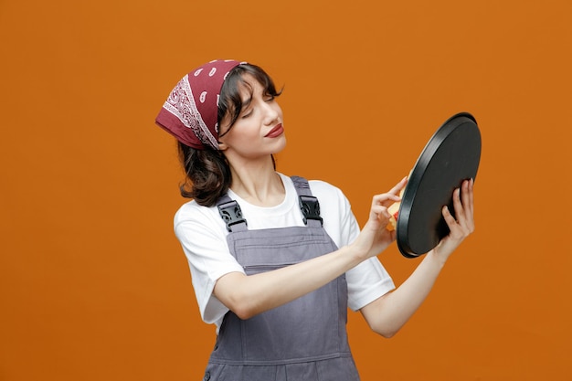 Concentrated young female cleaner wearing uniform and bandana cleaning tray with sponge isolated on orange background