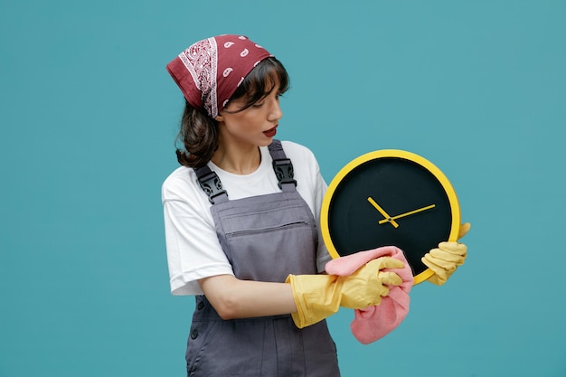 Concentrated young female cleaner wearing uniform bandana and rubber gloves holding clock wiping it with duster looking at it isolated on blue background