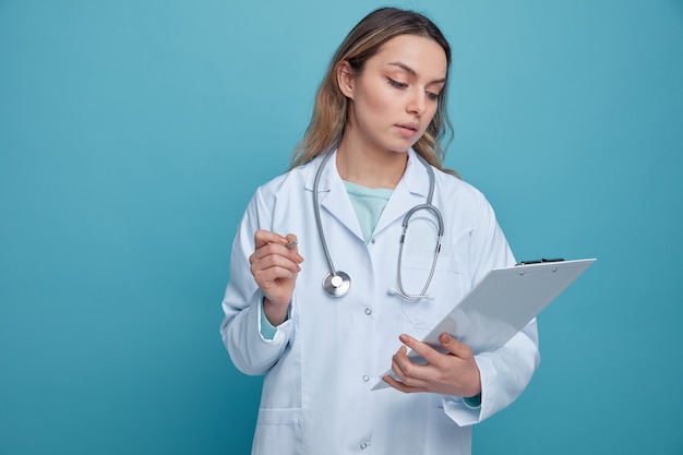 Concentrated young female doctor wearing medical robe and stethoscope around neck holding pen and clipboard looking at clipboard 