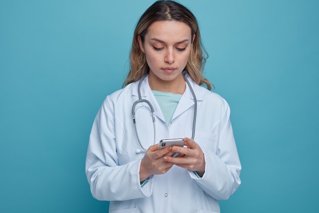 Concentrated young female doctor wearing medical robe and stethoscope around neck using her mobile phone 