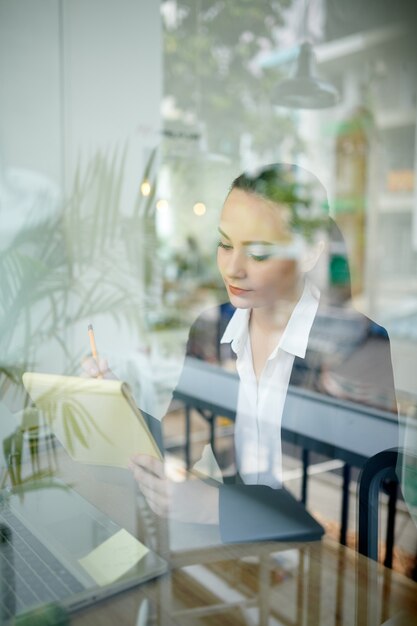 Concentrated young female entrepreneur reading and correcting document, view through the window