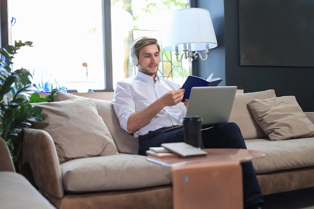 Concentrated young freelancer businessman sitting on sofa with laptop and examining documents