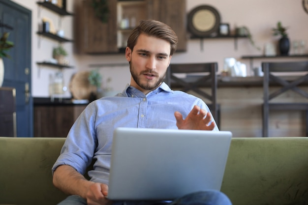 Concentrated young freelancer businessman sitting on sofa with laptop, working remotely online at home.