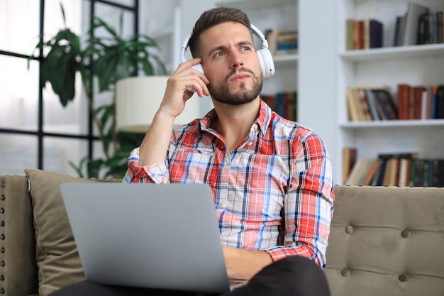 Concentrated young freelancer businessman sitting on sofa with laptop, working remotely online at home.