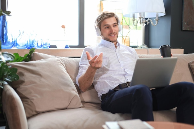 Concentrated young freelancer businessman sitting on sofa with laptop working remotely online at home