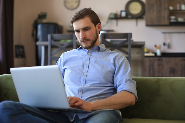 Concentrated young freelancer businessman sitting on sofa with laptop, working remotely online at home.