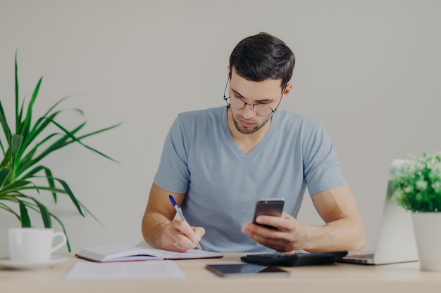 Concentrated young male accountant writes in notebook, makes business analysis