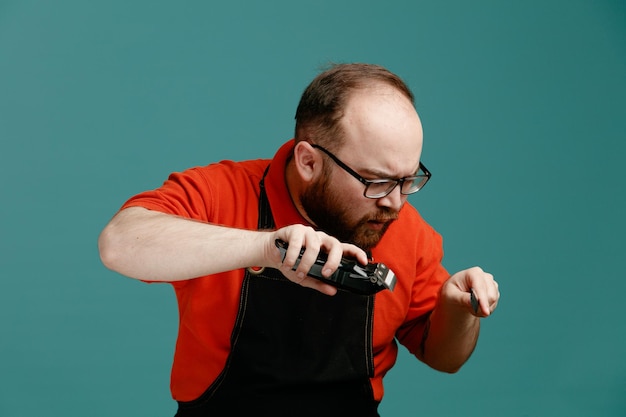 Concentrated young male barber wearing glasses red shirt and barber apron bending head down holding hair clipper and teaser comb in air isolated on blue background