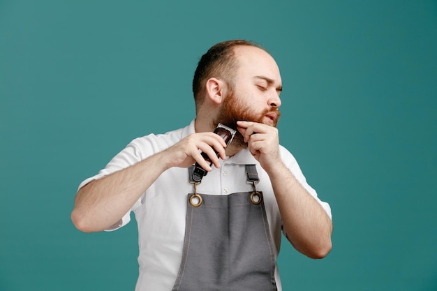 Concentrated young male barber wearing white shirt and barber apron turning head to side grabbing beard trimming his own beard with hair trimmer isolated on blue background