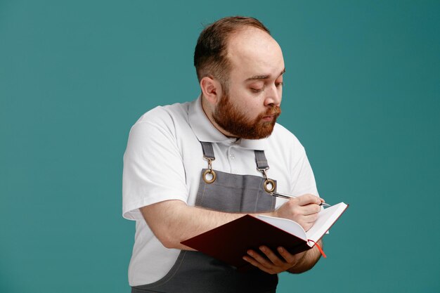 Giovane barbiere maschio concentrato che indossa una camicia bianca e un grembiule da barbiere che scrive con la penna sul blocco note isolato su sfondo blu