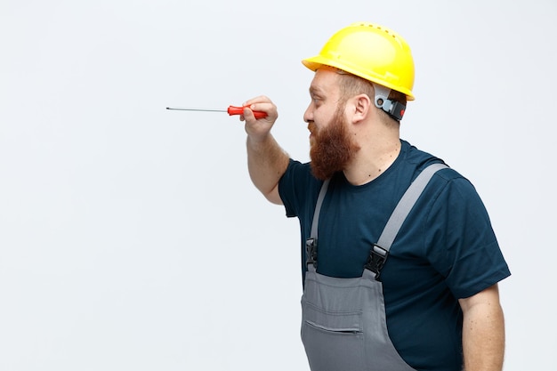 Concentrated young male construction worker wearing safety helmet and uniform standing in profile view using screwdriver looking at it isolated on white background with copy space