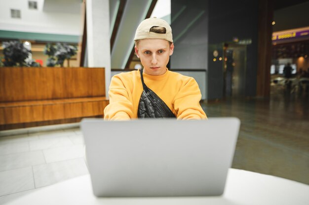 Concentrated young man in bright casual clothes uses a laptop in a mall cafe with a serious face