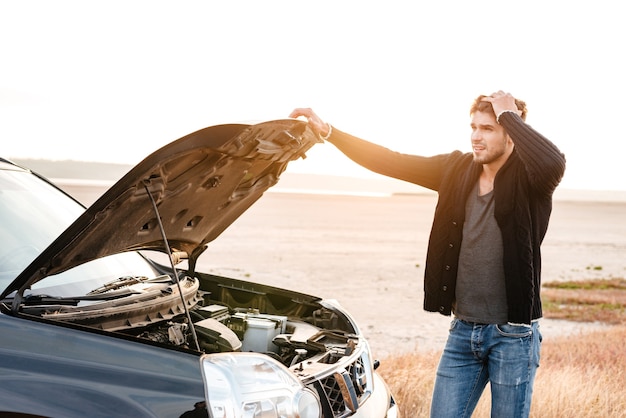 Concentrated young man holding hands on vehicle hood and looking inside it while standing outdoors