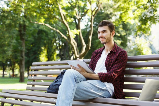 Concentrated young man making notes in workbook and using laptop in park. Technology, communication, education and remote working concept, copy space