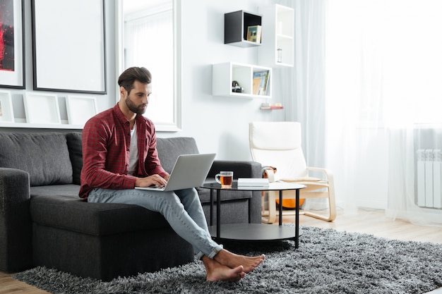 Photo concentrated young man sitting on sofa using laptop computer