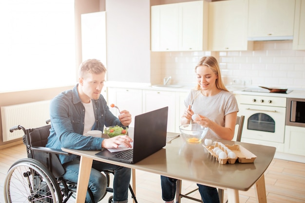 Concentrated young man on wheelchair working with laptop and eating salad. Studying with disability and inclusiveness. Guy with special needs. Young woman sit besides and cooking. Breaking eggs.