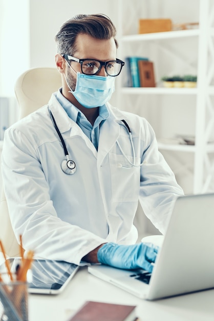 Concentrated young man in white lab coat and protective mask working using laptop while sitting indoors