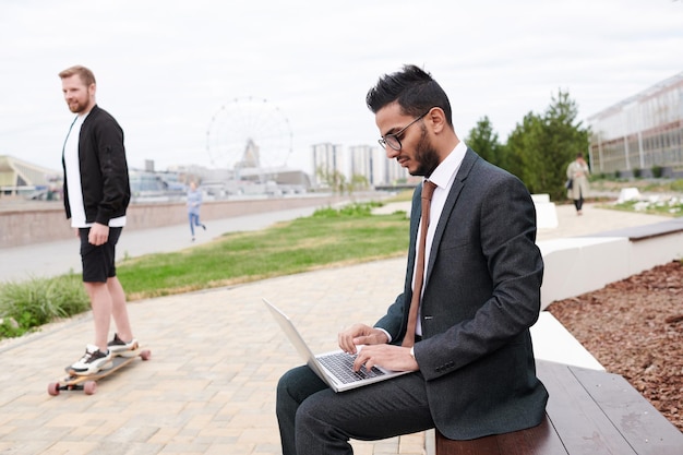 Concentrated young middleeastern manager in formal suit sitting on bench in city park and working with laptop