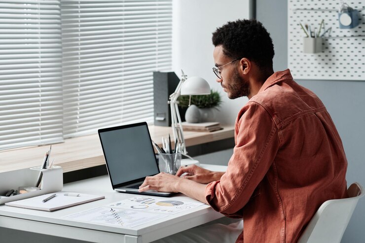 Photo concentrated young mixed race financial advisor in coral shirt sitting at desk and using laptop in o