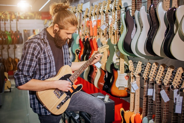 Concentrated young musician stand and play on electric guitar. He looks at it. Guy is in music shop. Young hipster concentrated on playing.