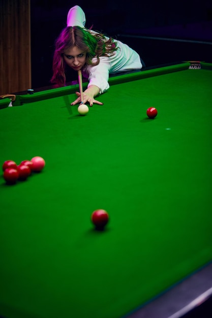 Concentrated young woman leaning on billiards table playing snooker game lifestyle magazine about