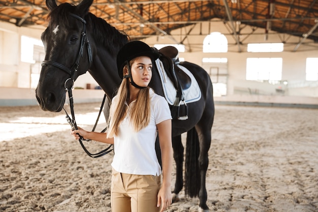 concentrated young woman wearing hat with horse in countryside in corral on training area.