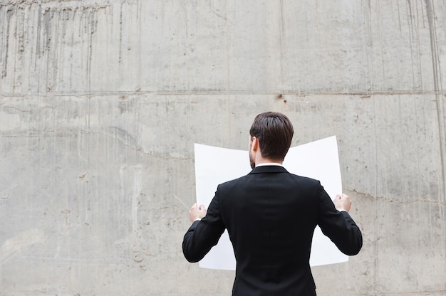 Concentration at work. Rear view of young man examining blueprint while standing outdoors and against the concrete wall