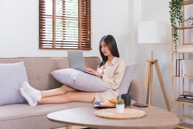 Concept of activity in living room Young Asian woman typing data on laptop to surfing social media