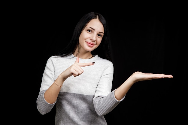 The concept of advertising. Positive, pretty, caucasion woman with a beaming smile and bright sweater on a black space indicates two index fingers and looks at the empty copy space.