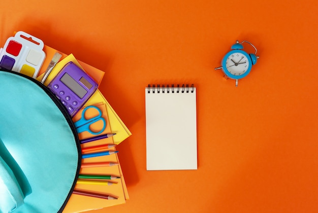 Concept back to school. Full turquoise School Backpack with supplies on orange. FLat lay.