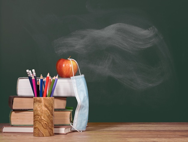 Concept for back to school with coronavirus or Covid19 with books and apple against a chalkboard with face mask