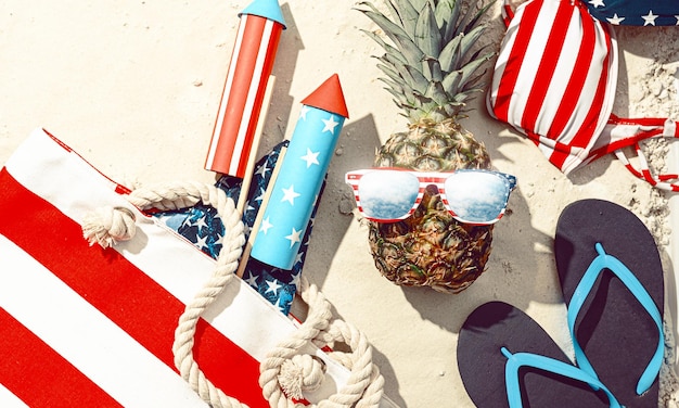 Concetto di festa in spiaggia in onore del giorno dell'indipendenza americana 4 luglio ananas con occhiali stilizzati come bandiera americana sulla sabbia sulla spiaggia