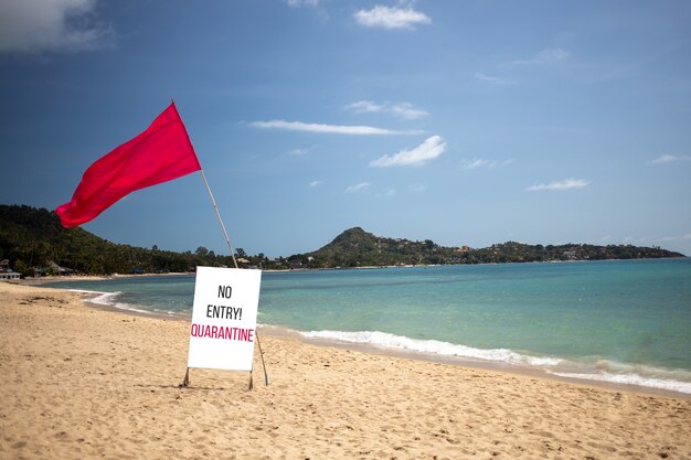 The concept of a closed beach, does not work, pandemic, the ban on travel and movement. there is a red flag and a sign that says "no entry, quarantine" on an empty tropical beach on a nice day.