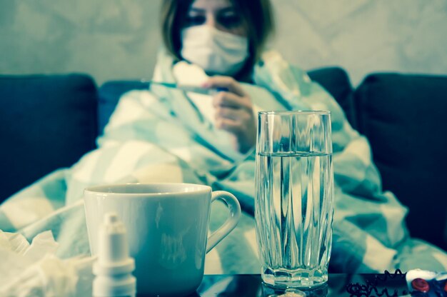 Il concetto del comune raffreddore o dell'influenza stagionale una giovane ragazza in maschera guarda un termometro seduto su un divano avvolto in una coperta vicino a un tavolo con medicinali beve una bevanda calda curativa quarantena