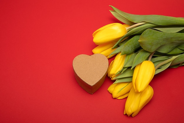 Concept of congratulations on holiday Mom's Day, Valentine's Day. Yellow tulips and heart-shaped gift box made of craft cardboard on red background. Copy space, mock up. Close up photo