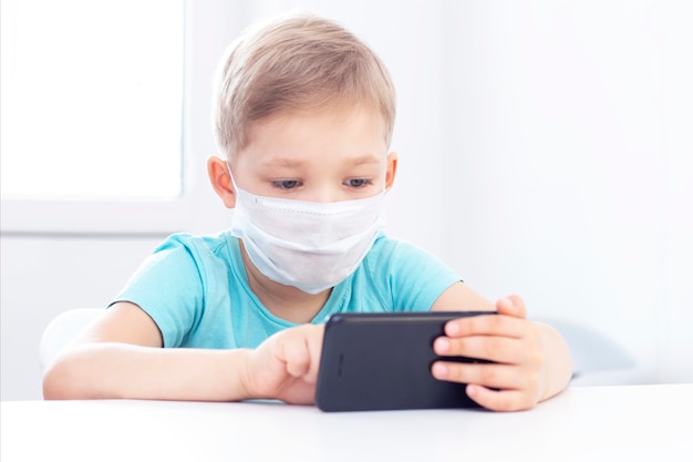 Concept of coronavirus infection. boy in a medical mask sits on a chair indoors and plays a gadget.