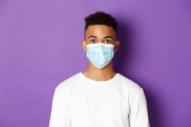 Concept of coronavirus, quarantine and social distancing. Close-up of young african-american guy in white sweatshirt and medical mask, looking at camera, standing over purple background.