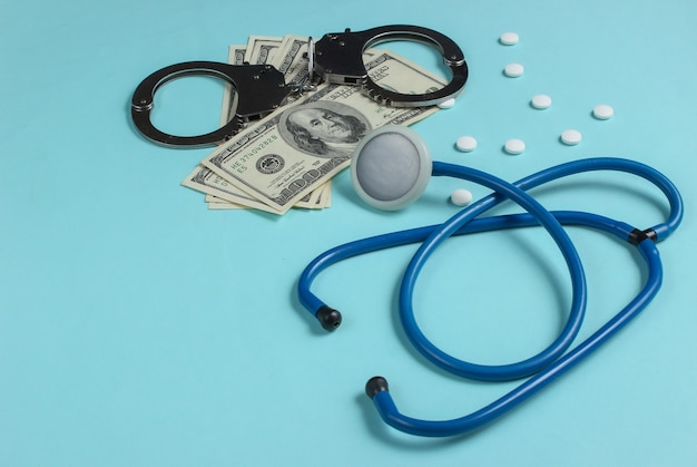 Photo concept of corruption in medicine. stethoscope, pills and handcuffs with hundred dollar bills on blue background. medical still life. punishment for crime