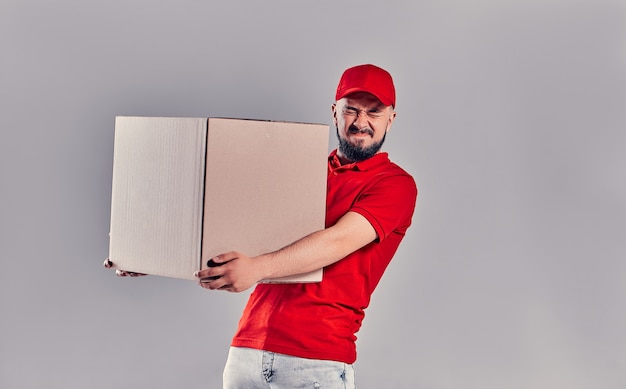 Концепция курьерской и курьерской службы. Гримасничающий красивый парень пытается держать в руке большую картонную коробку, изолированную на сером фоне.