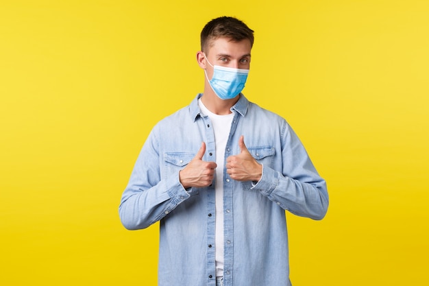 Concept of covid-19 pandemic outbreak, lifestyle during coronavirus social distancing. Confident handsome blond guy in medical mask and casual outfit showing thumbs-up over yellow background.