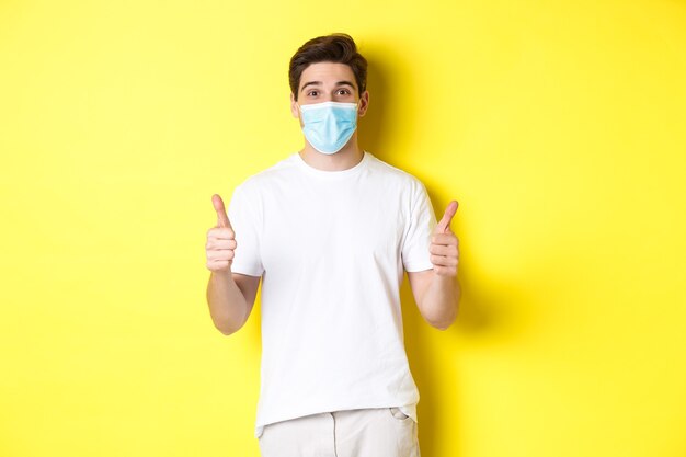 Concept of covid-19, quarantine and lifestyle. Happy man in medical mask showing thumbs up, approve or saying yes, like something good, yellow background