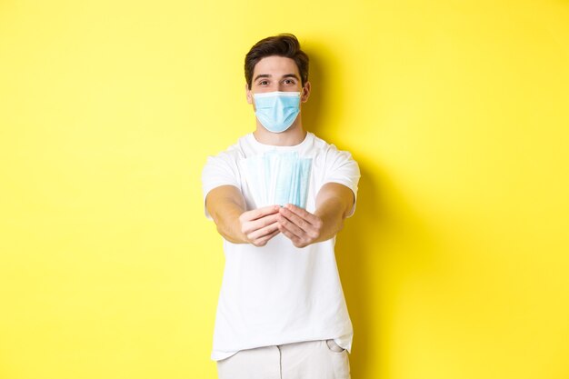 Concept of covid-19, quarantine and preventive measures. Young caucasian man giving medical masks for you, standing against yellow background