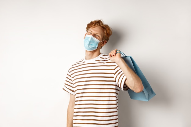 Concept of covid-19 and shopping. Satisfied young man looking pleased after shopping, wearing face mask, holding paper bag and smiling, white background.