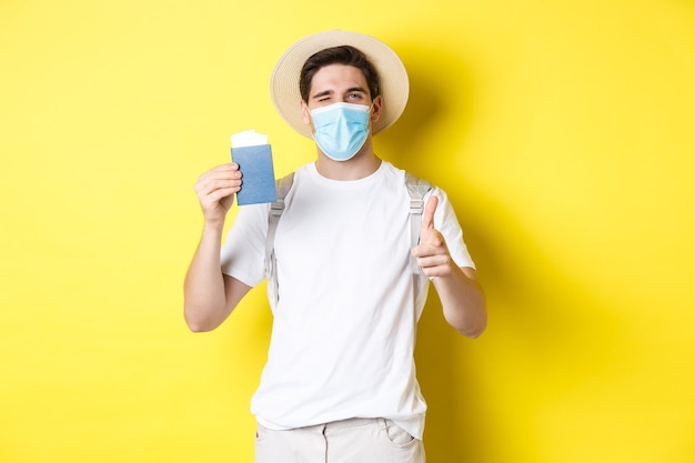 Concept of covid-19, tourism and pandemic. Happy guy tourist in medical mask showing passport with tickets for vacation, winking and pointing at camera, yellow background