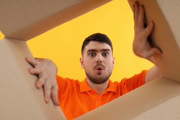 Photo concept of delivery gift surprise young man look in to cardboard box on yellow background
