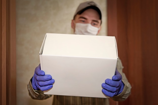 2019年検疫中の配達品の概念-ncov。医療マスクと青い手袋の若い男は、手に段ボール箱を保持します。非接触型配信ウイルス保護、デザインまたはボックス上のテキスト用の空きスペース