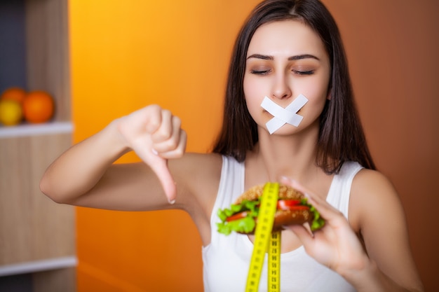 Concept of diet cute woman with sealed mouth keeps greasy burger